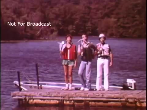 The still features three individuals standing on a wooden dock by a body of water, all wearing life jackets. They appear to be cheerfully singing and promoting boating safety. The background shows trees reflecting off the water, creating a serene outdoor setting. The image is clearly marked as “Not For Broadcast,” indicating it is part of a public service announcement.
