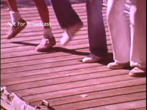 The still shows a group of people on a wooden dock, focusing on their feet as they participate in a coordinated activity, likely singing or dancing. The scene emphasizes a playful and engaging atmosphere, aligned with the PSA's message about safe boating practices. The setting suggests a casual, outdoor environment, enhancing the friendly and inviting tone of the public service announcement.