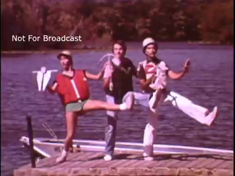 The still depicts three individuals on a dock by a body of water, performing a lively song and dance for a public service announcement about safe boating. One person wears a life jacket and shorts, while another is dressed in a casual outfit, and the third wears a hat. They enthusiastically strike poses and kick their legs, promoting boating safety in a fun and engaging manner. The background shows a boat and trees, adding to the outdoor setting. The image is marked 'Not For Broadcast.'