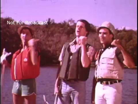 The still from the 'Singing National Safe Boating Council PSA' features three individuals near water, promoting safe boating practices. They appear engaged and cheerful, likely singing or performing a routine. The individuals are wearing life jackets, emphasizing boat safety. The background suggests a natural setting, reinforcing the outdoor theme of boating safety. The overall tone is upbeat and instructional.
