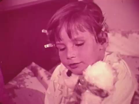 The still from the 1969 Easter Seals PSA features a young child with short hair, sitting against a softly colored background. The child appears to be introspective, holding a stuffed animal close. The scene conveys a gentle, caring atmosphere, typical of public service announcements aimed at raising awareness for disabilities and support through Easter Seals. The visual style is characterized by a warm, pinkish hue, giving it a nostalgic feel.