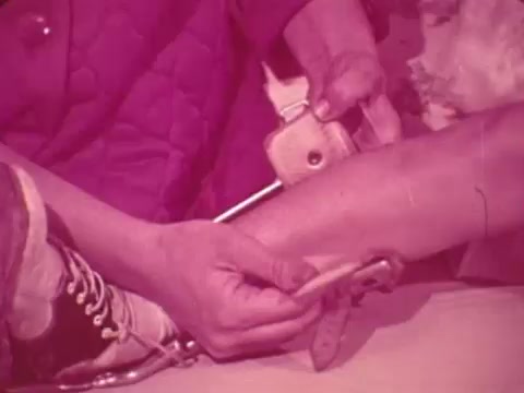 The still from the 1969 Easter Seals PSA shows a close-up of hands fitting a brace or support device onto a person's leg. The focus emphasizes the care and attention involved in assisting individuals with disabilities. The color tone is a pink hue, creating a soft yet impactful visual that highlights the importance of support and rehabilitation in disability services.