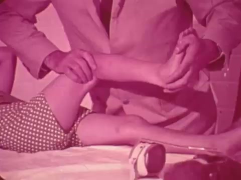 The still depicts a man in a suit gently examining the leg of a child, who is likely receiving physical therapy or a medical assessment. The child's leg is bare, and they are wearing polka-dotted shorts. The scene has a reddish tint, typical of older film stock, emphasizing a focused and caring interaction, likely aimed at promoting awareness of services provided by Easter Seals for children with disabilities.