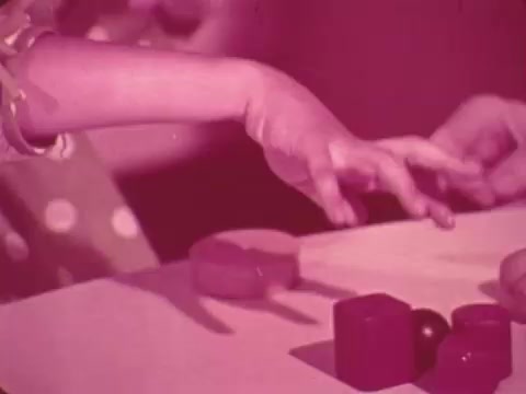 The still from the 1969 Easter Seals PSA features a close-up of children's hands interacting with colorful geometric blocks on a table. The image has a distinctive pinkish hue, emphasizing the focus on the children's actions, which may highlight themes of learning, play, and developmental support. The scene suggests engagement and participation, aligning with the message of Easter Seals in promoting services for children with disabilities.