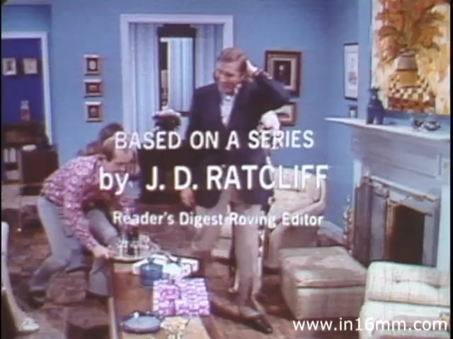 The still from 'I Am Joe's Stomach' features a scene with two characters in a domestic setting that reflects the 1980s aesthetic. One character appears to be adjusting his hair while another is interacting with a table filled with items, likely in a humorous or awkward situation. The background adds to the retro vibe, with patterned wallpaper and vintage furnishings. The text at the top indicates that the piece is based on a series by J.D. Ratcliff, suggesting it aims to impart knowledge about human health, specifically the stomach and its functions, through engaging storytelling and animation.