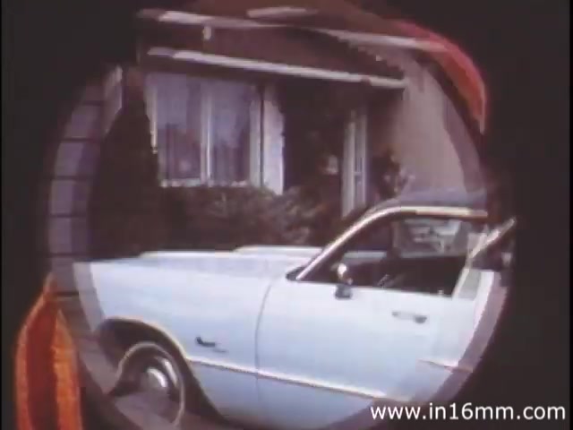The still from 'I Am Joe's Stomach' (1980s version) showcases a 16mm film aesthetic with a circular framing device. The image captures a scene outside a house, focusing on a light blue car parked prominently in the foreground. The surrounding environment includes what appears to be well-kept shrubs and a window in the background, hinting at domestic life. This visual context ties into the film's exploration of human stomach functions and health, using both animation and live action to depict the bodily experience. Overall, the still captures a moment that connects the everyday with the narrative of personal health issues.
