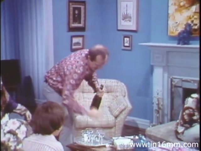 The still from 'I Am Joe's Stomach' (1980s version) features a vibrant living room scene where a man, dressed in a patterned shirt, is seen excitedly popping open a bottle of champagne. He stands near a glass coffee table adorned with small glasses, with a casual yet lively demeanor. In the foreground, a seated figure appears to be engaged in the celebration, while various framed artworks adorn the blue walls, contributing to a cozy, domestic atmosphere. This scene juxtaposes live action with the film's underlying theme of exploring human stomach functions and gastrointestinal health through relatable experiences.