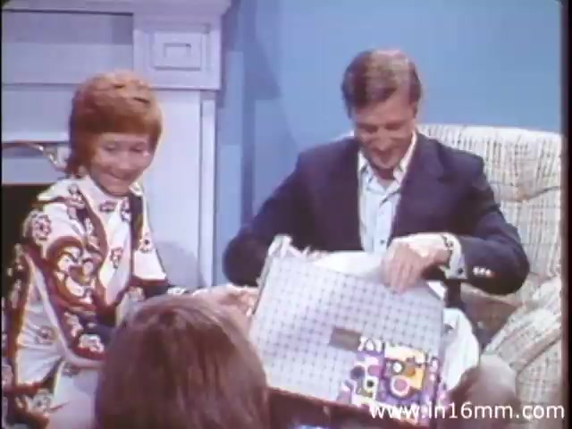 The still from 'I Am Joe's Stomach,' a 1980s production by Reader's Digest Association, features a lively scene where a man is opening a gift, showcasing expressions of excitement and joy. Sitting next to him is a woman, possibly sharing in the moment, illustrating a warm and friendly atmosphere. The setting is casual, with a cozy living room backdrop, providing a relatable context. This segment likely serves as a contrast to the film's educational content on digestive health, using personal narratives to engage viewers in understanding the function and care of the human stomach.