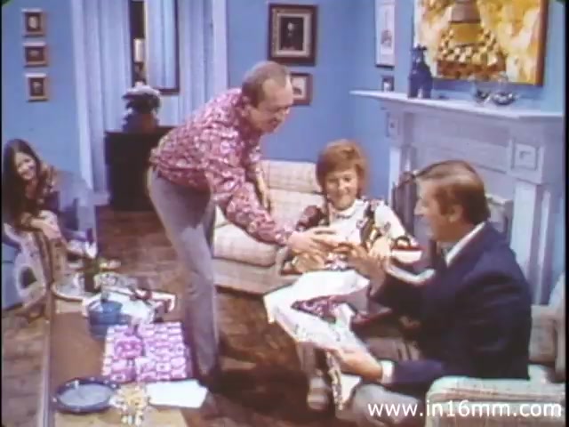 The still from 'I Am Joe's Stomach' features a scene set in a living room, characterized by its 1980s decor. The main characters include a man in a brightly patterned shirt, who is interacting with a seated woman and another man dressed in a suit. They appear engaged in conversation, possibly discussing food or digestion, reflecting the film's focus on the human stomach and its functions. Surrounding them are elements typical of family gatherings, such as snacks and drinks, enhancing the casual, domestic atmosphere. The animation and live-action juxtapose educational content with relatable human experiences regarding stomach health.