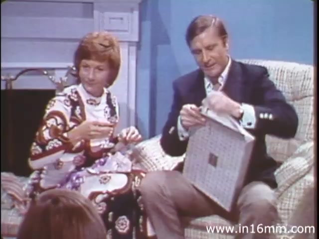 The still from 'I Am Joe's Stomach' features a man and a woman seated on a patterned couch in a domestic setting. The woman, dressed in a colorful, patterned dress, appears engaged, while the man is unwrapping a gift, possibly depicting a lighthearted moment. The scene exemplifies the film's educational approach, combining live sequences with animation to emphasize the functioning of the human stomach and related health issues in a relatable manner.