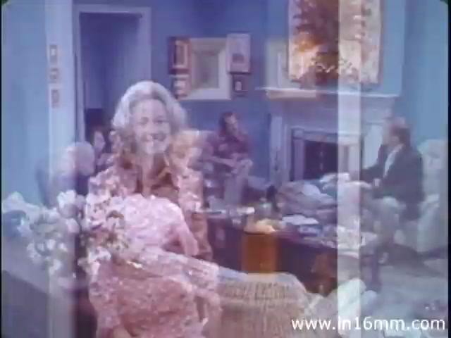 The 16mm still from 'I Am Joe's Stomach' captures a scene featuring a woman smiling in the foreground, with a living room setting in the background. The room appears to be lively, filled with people engaged in conversation. This educational film blends live-action sequences with animation to illustrate the functions of the stomach, highlighting digestive health through the narrative of a character facing digestive issues.