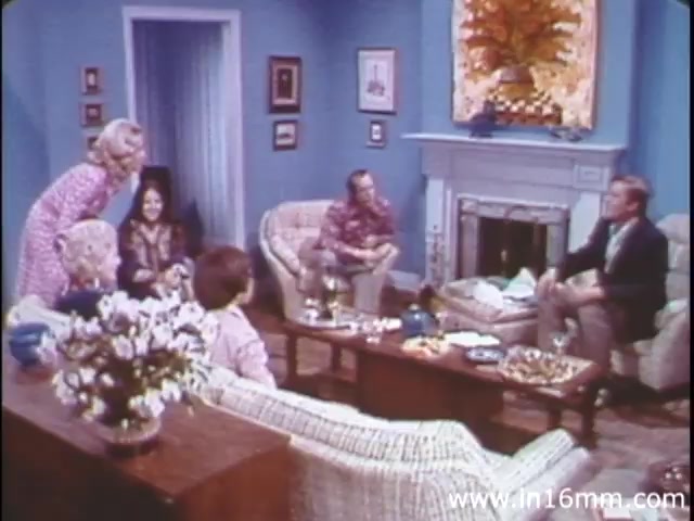 The still from 'I Am Joe's Stomach' features a group of people gathered in a cozy living room, typical of the 1980s, with light blue walls and a floral painting. The scene includes six individuals: a woman in a pink dress leaning over to speak with another woman, while a man in a patterned shirt sits nearby. An additional man in a blazer appears to be leading the conversation. The setting suggests a casual, educational discussion about digestive health, mirroring the film's focus on the human stomach and its functions through relatable narratives.