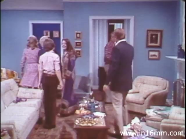 The still from 'I Am Joe's Stomach' features a living room setting typical of the 1980s, characterized by pastel blue walls and a casual, homey atmosphere. A group of people is present, engaged in conversation or interaction, conveying a sense of camaraderie. The scene is cluttered with various items, hinting at a casual gathering or party. The film uses this relatable environment to introduce its educational content about the human stomach and digestion through engaging storytelling, showcasing both animation and live-action.