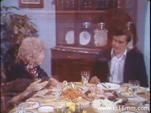 The still from 'I Am Joe's Stomach' features a scene likely set at a dining table, capturing a moment of conversation between two characters: a woman with curly hair and a man dressed in a suit. They appear engaged in a discussion, surrounded by a meal that includes various dishes and drinks. The background includes a cabinet with glassware, suggesting a domestic setting. This live-action sequence complements the film's educational theme about the human stomach's functions and health, illustrating the social context of eating while hinting at underlying digestive issues.