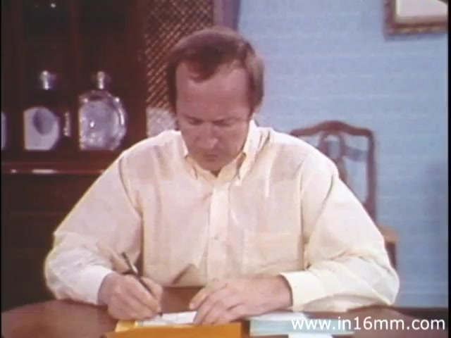 The still from 'I Am Joe's Stomach' features a man in a light-colored shirt sitting at a table, focused on writing something down. The background includes a cabinet with items displayed and a chair nearby, creating a homey, domestic setting. The film employs a blend of animation and live action to illustrate the human digestive system, specifically addressing issues related to stomach health in a relatable and engaging manner. The context highlights the interactions between a person and the functioning of their stomach, ultimately emphasizing the importance of digestive care.