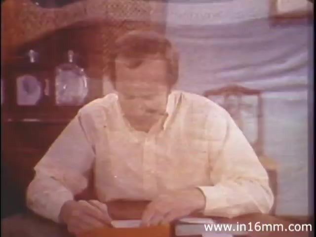 The still from 'I Am Joe's Stomach' features a man seated at a table, appearing to write or draw. He looks focused, with his head slightly bent and a serious expression. The background includes an assortment of items, hinting at a domestic setting. This educational film uses a mix of animation and live action to explore the functions of the human stomach, emphasizing its care through relatable experiences. The concept draws upon the metaphor of 'Joe,' personifying the stomach's needs and challenges.