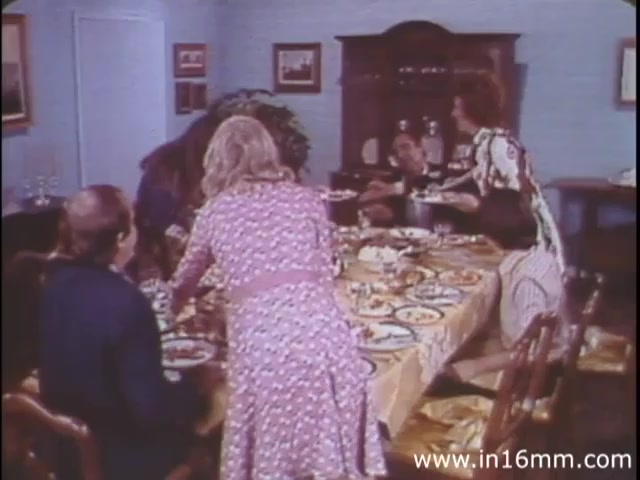 The still from 'I Am Joe's Stomach' depicts a lively dinner scene, showcasing a group of people gathered around a table laden with various dishes. The setting is a homey dining room, with a warm, inviting atmosphere created by the colorful decor and casual interactions. A woman serves food, hinting at themes of overeating and digestive issues, which are central to the narrative. The use of live-action highlights the relatable experiences of the characters, embodying the struggles of digestion in a lighthearted manner typical of the 1980s approach to health education.