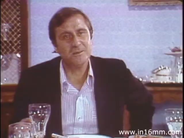 The still from 'I Am Joe's Stomach' depicts a middle-aged man seated at a dining table, dressed in a black suit jacket over a light-colored striped shirt. He appears to be in mid-conversation, suggesting a humorous or informative discussion about the intricacies of stomach function. The background features a dining setting, indicating a casual atmosphere where health topics are approached in a light-hearted manner. The film employs a mix of live-action and animation to illustrate the workings of human digestion, focusing on the consequences of poor dietary choices.