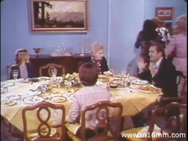 The still from 'I Am Joe's Stomach' features a family scene around a dining table set for a meal. The table is adorned with a yellow tablecloth and appears to have various dishes laid out. A man, animatedly speaking, is seated at the table, while others, including children, appear engaged in conversation. The background shows a painted landscape on the wall and people moving around the dining area, capturing a sense of familial interaction. This context highlights the film's focus on the functions of the human stomach and the importance of food choices, illustrating the narrative through this relatable family setting.