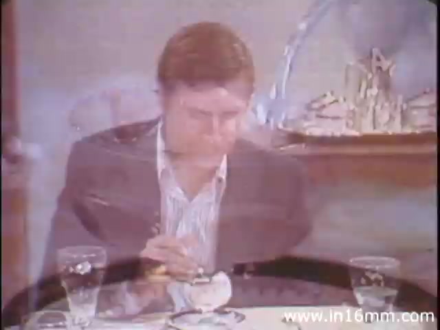 The still from 'I Am Joe's Stomach' features a man seated at a dining table with two glasses of water in front of him. He is engaged in eating, appearing contemplative or concerned, which aligns with the film's theme of exploring the functions and care of the human stomach. The animated sequences interspersed with live-action highlight the intricacies of digestion, emphasizing the importance of food choices and stomach health through the character's experiences. The overall tone balances education with personal narrative, typical of the 1980s documentary style.