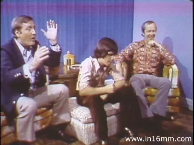 The still from 'I Am Joe's Stomach,' a 1980s educational film by Reader's Digest, features three men seated on a couch in a casual set. The man on the left animatedly gestures with his hands, likely discussing a topic related to the stomach's functions. The man in the center appears more subdued, possibly contemplating or reacting to the conversation. The third man on the right, with a patterned shirt, has a relaxed demeanor, sharing the moment. The backdrop is a simple blue, enhancing the informal atmosphere. This animated educational piece combines live action and animation to explore stomach health through engaging narratives.