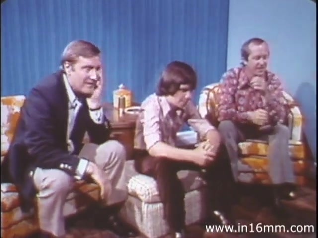 The still from 'I Am Joe's Stomach' features three men seated in a living room, engaged in what appears to be a serious discussion. The setting includes retro furniture with a patterned couch and brightly colored walls typical of the 1980s aesthetic. The individuals express varying degrees of concern, reflecting the educational and possibly humorous tone of the film, which uses a mix of animation and live action to explore digestive health in an accessible way. The context suggests they are likely reacting to Joe's stomach-related issues, emphasizing the importance of digestive care.