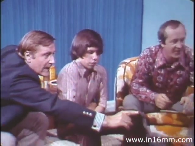 The still from 'I am Joe's Stomach' features three characters seated in a living room setting. The men are engaged, with one reaching forward to point at something, suggesting an animated discussion. The backdrop consists of a blue curtain, and the furniture, including an orange and yellow patterned couch, is indicative of the 1970s aesthetic. This scene likely serves to bridge live action with animated sequences, emphasizing the film's educational approach to explaining the human stomach's functions and care through relatable, everyday experiences.