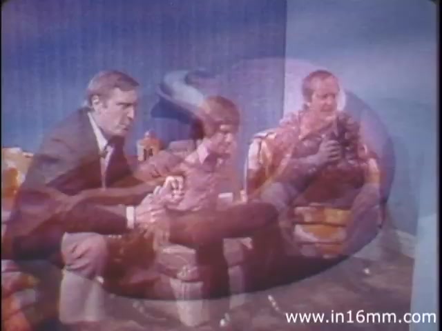 The still from 'I Am Joe's Stomach' features three men sitting on couches, engaged in conversation. The scene utilizes three-dimensional animation layered over live-action, illustrating the complexities of stomach function. The men appear to represent different perspectives or experiences related to digestive health, emphasizing the importance of proper care. The overall aesthetic reflects the 1980s style, combining educational content with a personable narrative approach.