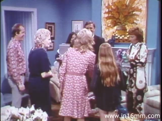 In the 16mm still from 'I Am Joe's Stomach,' a lively scene unfolds in a well-decorated living room, indicative of the 1980s style. The focus is on a group of people interacting, showcasing both adults and children dressed in vintage clothing. The background features bright colors and decorative elements typical of the era. This segment likely serves as a narrative bridge, integrating live-action with the educational content that explains the functions of the stomach through relatable human experiences. The overall tone is light-hearted, reflecting the film's intention to engage and inform viewers about health in a relatable manner.