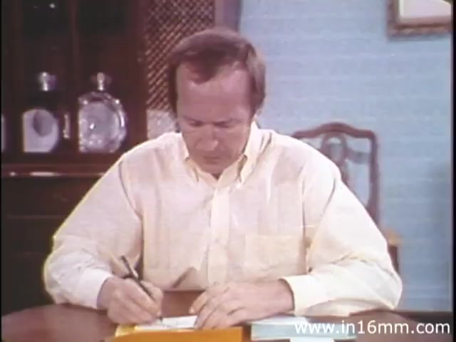 The still from 'I Am Joe's Stomach' depicts a man seated at a table, engaged in writing. He appears focused, wearing a light-colored button-up shirt. The setting features a vintage décor, with a wooden table and items in the background, including a decorative piece. The film, produced in the 1980s by Reader's Digest Association, employs a mix of animation and live-action to illustrate the functions and care of the human stomach through a narrative centered on digestive health issues.