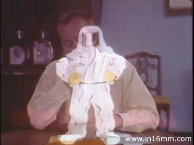 The still depicts a scene from the 1980s educational film 'I Am Joe's Stomach,' produced by Reader's Digest Association. It features a humorous and educational approach, combining three-dimensional animation with live-action sequences. In this moment, a character seated at a table seems to be interacting with an animated representation of a stomach, illustrated in a whimsical manner. The figure is engaging and stylized, emphasizing the intricacies of stomach functions. This visual style serves to engage viewers in understanding the importance of digestive health through relatable and entertaining narratives.