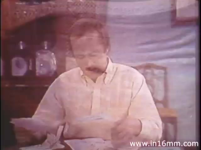 The still from *I Am Joe's Stomach* depicts a man sitting at a table, seemingly engaged in reading or reviewing documents. He has a mustache and wears a light-colored shirt. The background includes decorative elements, likely from the 1980s, such as an ornate wooden structure and glass bottles. The scene reflects the film's educational approach, utilizing a blend of live-action and animation to explore human stomach functions and the importance of digestive health, framed through the man's experiences with gastrointestinal issues.