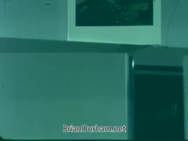 The still from the 'Point Park College PSA' features a dimly lit room with a predominantly greenish hue. In the background, a framed photo is slightly visible, while various surfaces, possibly a desk or shelf, are in focus. The overall composition conveys a sense of academic or promotional context, typical for a public service announcement about the college.