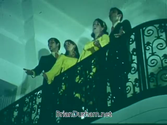 The still features four individuals performing on a balcony, dressed in formal attire. Two are wearing black suits while the other two are in bright yellow dresses. They appear to be singing or expressing themselves dramatically, with visible enthusiasm and engagement. The lighting casts a vibrant green hue over the scene, creating a visually striking atmosphere. The backdrop includes an ornate, decorative railing.
