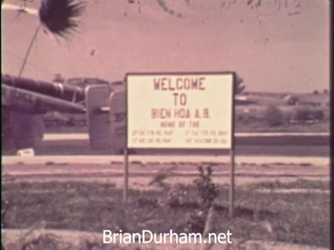The still features a sign stating 'WELCOME TO BIEN HOA A.B.' with a backdrop that includes military aircraft. The setting is likely a U.S. military base in Vietnam during the war era, conveying a sense of military presence and the context of that time. The visual serves to connect viewers with the historical significance of the Vietnam War and the initiative to promote savings bonds as part of support for the troops.