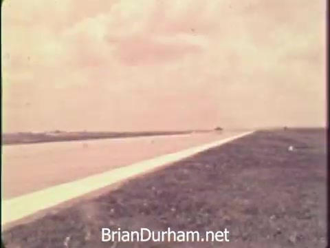 The still depicts an expansive, open landscape featuring a wide road that stretches into the distance. The sky is light, hinting at either an overcast day or nostalgic tone with pastel colors. This setting evokes a sense of tranquility amidst the chaotic backdrop of the Vietnam War era, aligning with the theme of encouraging savings bonds to support the troops. The overall composition conveys both a sense of journey and the drive for unity during challenging times.