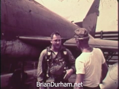The still from the 1968 PSA depicts two men near an aircraft. One man, possibly a soldier, is wearing a flight suit and harness, suggesting readiness for flight or military action. The other man, dressed casually in a white shirt and cap, appears to be engaged in conversation with the soldier. The setting indicates a military environment, likely related to the Vietnam War, underscoring themes of patriotism and support for the war effort through savings bonds. The overall tone reflects the era's military focus.