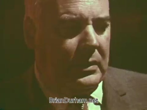 The still shows a middle-aged man with a serious expression, likely delivering a message or speech. He is dressed in a suit, and the lighting creates a dramatic effect, emphasizing his facial features. The context suggests he is involved in a public service announcement related to Boy Scouts, focusing on themes of responsibility or character development.