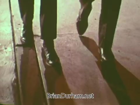 The still features a close-up of two pairs of feet walking on a wooden surface, likely in a public setting. The footwear is polished, suggesting a formal or purposeful context. The lighting creates shadows, adding depth to the scene. The overall tone is reminiscent of a mid-20th-century public service announcement related to the Boy Scouts, emphasizing themes of duty and camaraderie.