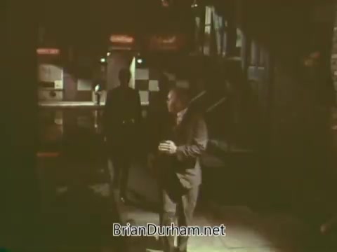 The still is from a 1960s Boy Scouts PSA, depicting a scene with a somber atmosphere. Two figures, one in the foreground and another in the background, are shown in a dimly lit environment, possibly a street or urban setting. The overall tone suggests a serious or cautionary message, typical of public service announcements from that era.