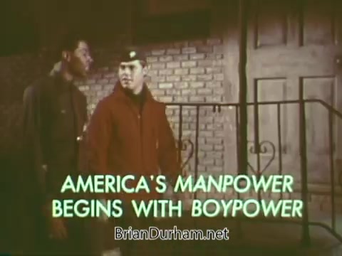 The still from the 'Boy Scouts PSA - 1960's' features two young boys, one dressed in a scout uniform, standing together in front of a brick wall and a staircase. The screen prominently displays the text, 'AMERICA'S MANPOWER BEGINS WITH BOYPOWER,' emphasizing a message about the importance of youth and scouting in building future strength. The overall tone suggests camaraderie and teamwork.