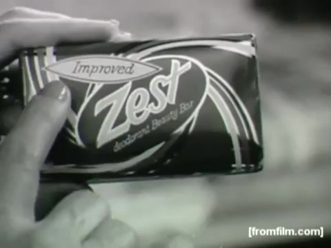 The still features a hand holding a bar of 'Improved Zest' deodorant beauty soap. The packaging is sleek with bold typography and swirls, emphasizing its 'improved' formula. The image is in black and white, characteristic of 1960s commercials, highlighting the product's appeal and modernity for the era.