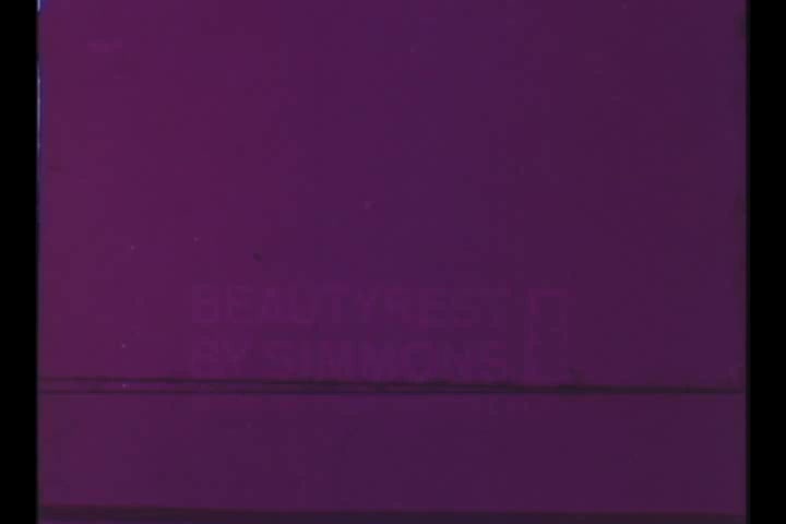 The still from the 'Adorn Hair Spray' 1960's TV commercial features a purple-toned background with text partially visible, reading 'BEAUTY' and 'BY SIMMONS.' The image likely reflects the vintage aesthetic of the era, showcasing the product's branding and appeal. Overall, it captures a nostalgic look at mid-20th century advertising.