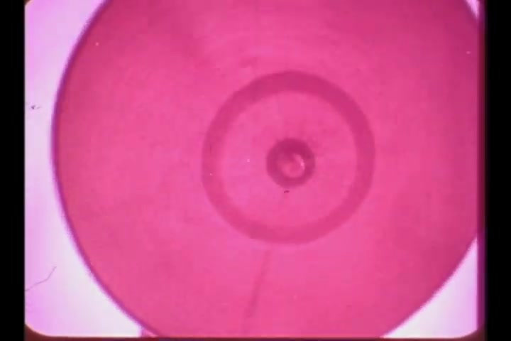 The still is a close-up view of a circular object, possibly a container or spray nozzle, rendered in a faded pink hue typical of aged 16mm film. The circular design features concentric rings and a central point, creating a visually intriguing pattern. The overall appearance suggests a vintage aesthetic, appropriate for a 1960s television commercial for Adorn Hair Spray.