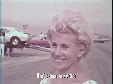 The still features a smiling Shirley Shahan, a professional drag racer, highlighting her confidence and positivity about the sport. She stands in a racetrack setting, with cars in the background, conveying an atmosphere of competition and excitement. Her expression reflects enthusiasm and assurance, aligning with her message about safety in drag racing, especially for women.