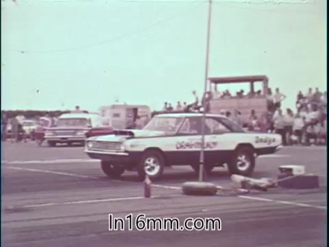 The still features Shirley Shahan's drag racing car, which is prominently positioned on a drag strip. In the background, a crowd watches, with several vehicles and a timing tower visible, suggesting a bustling event atmosphere. The focus is on the car, highlighting its stocker design, which is associated with Shahan's emphasis on safety in her racing narrative. The image encapsulates the blend of competitive spirit and safety ethos she promotes.