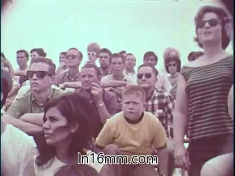 The still features a crowd watching a drag racing event, with a mix of spectators including men and women. The audience appears engaged, possibly reacting to the race or commentary. The atmosphere seems electric, with some individuals wearing sunglasses and casual clothing typical of the era. This scene emphasizes the excitement and community surrounding drag racing in the 1960s, which is complemented by Shirley Shahan's spoken emphasis on safety in the sport.