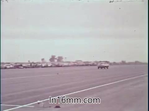 The still depicts a drag racing scene featuring a car speeding down a straight racetrack, with a row of parked vehicles in the background. The atmosphere suggests a bustling motorsport event, and the visual context aligns with the voice-over highlighting Shirley Shahan's expertise as a professional driver who prioritizes safety in her sport. The tone is confident and reinforces the notion that drag racing can be a safe pursuit for women.