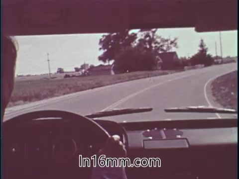 The still from the PSA features an interior view of a car with a driver's hand on the steering wheel. The focus is on a winding road ahead, showcasing a rural landscape with fields and trees in the background. The context highlights Shirley Shahan's expertise in drag racing and her emphasis on safety, challenging the perception of the sport as dangerous for women.
