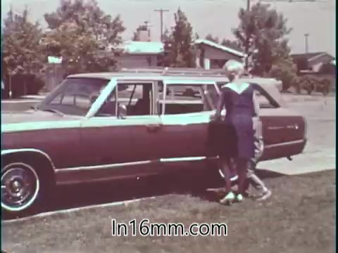 In the still from the 1960s PSA featuring Shirley Shahan, a professional drag racer, we see her positioned near a vintage station wagon, likely preparing for a car ride. The environment appears suburban, with homes and trees in the background. Shahan, dressed in a stylish outfit, embodies confidence and safety, reflecting her sentiment that drag racing can be safe for women. The image captures a moment that emphasizes both her identity as a driver and the importance of automotive safety.